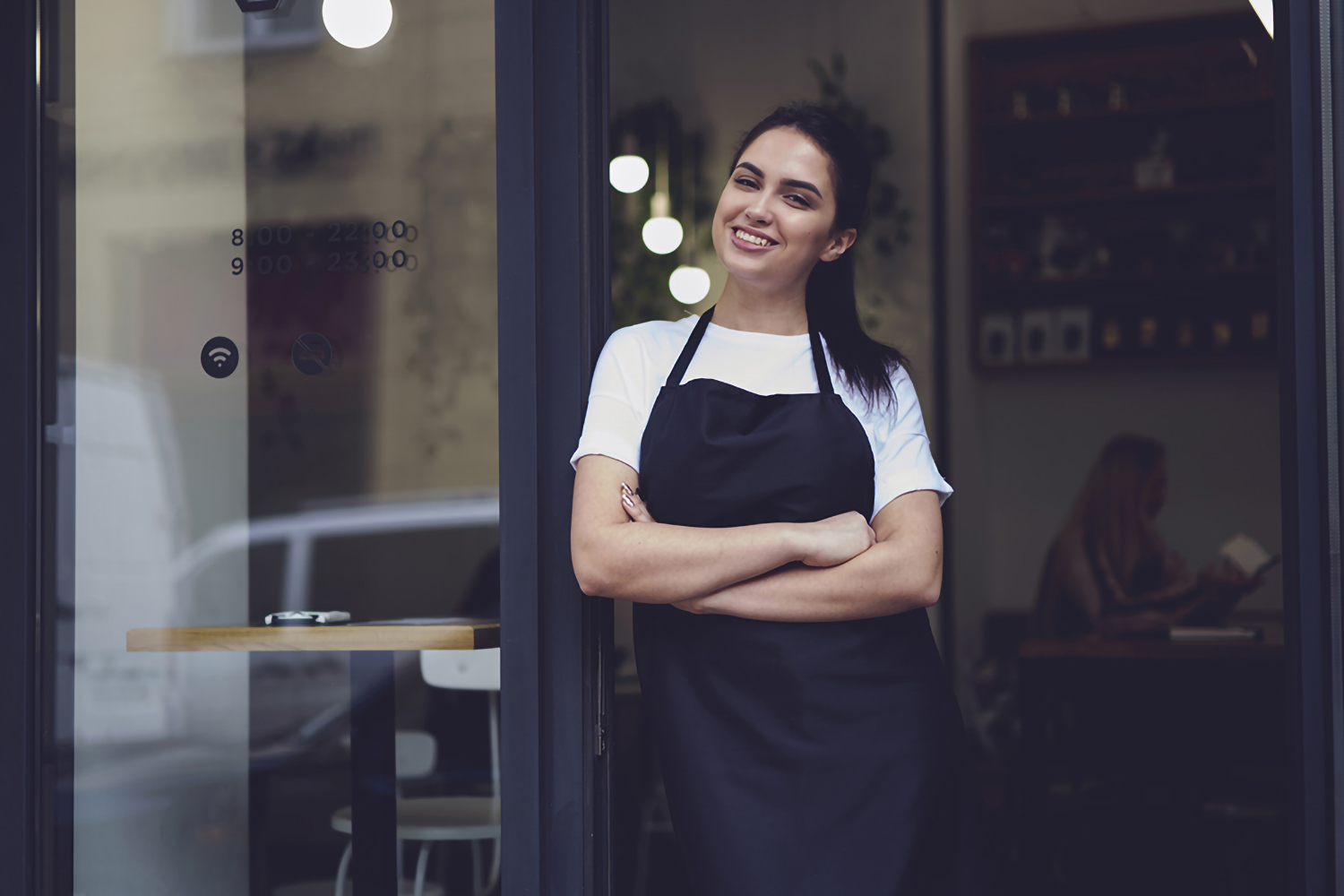 Half length portrait of young business woman waitress in black apron ready to attend new customers in her just opened coffee shop.Female owner of restaurant with smile standing near cafe door entrance