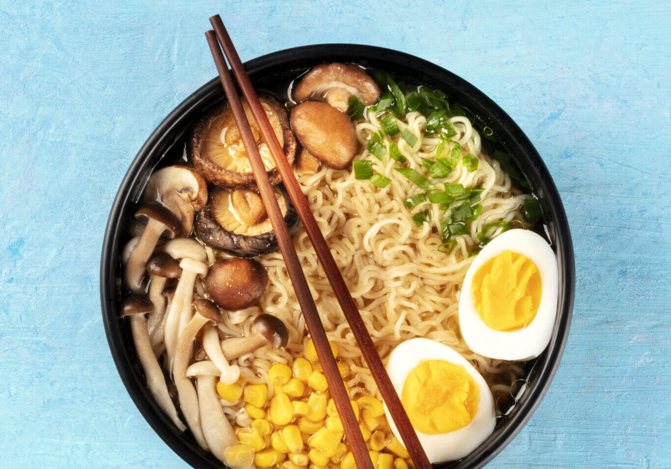Ramen. Soba noodles with eggs, shiitake mushrooms, and vegetables, top-down shot on a blue background with chopsticks and a place for text