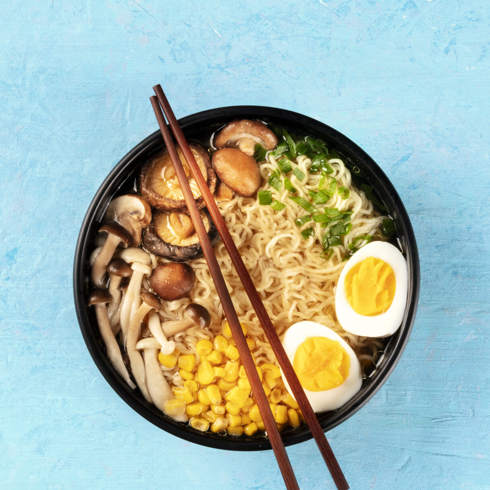 Ramen. Soba noodles with eggs, shiitake mushrooms, and vegetables, top-down shot on a blue background with chopsticks and a place for text